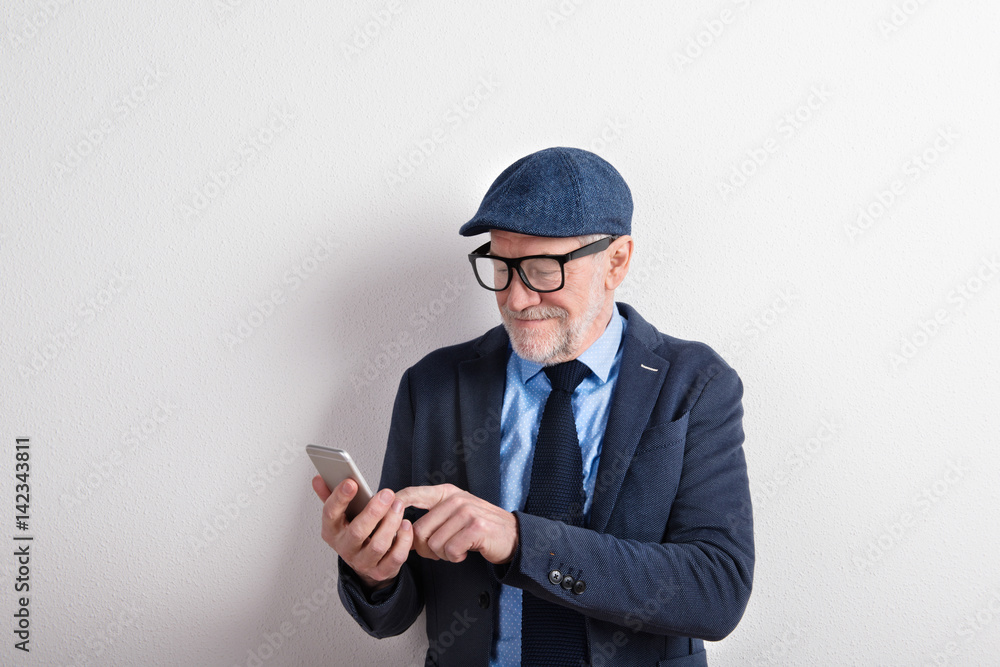 Smart senior man in suit, eyeglasses and cap, holding smartphone Smart senior man in suit, eyeglasses and cap, holding smartphone