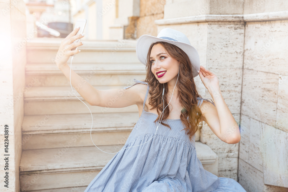 Fototapeta premium Smiling woman in hat making selfie with smartphone outdoors