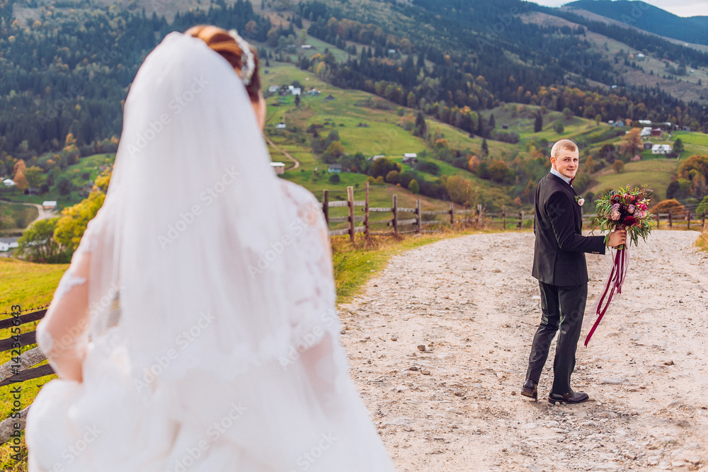 Bride and groom on gorgeous view of mountains,country road, sky, clouds on sunset. Newlyweds are after wedding ceremony. Bride is dressed in classic white wedding dress and bridal veil.