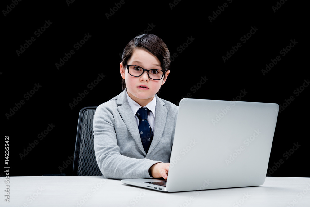 Confused little boy in formal suit and eyeglasses working with laptop Confused little boy in formal suit and eyeglasses working with laptop