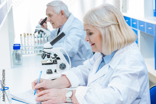 senior chemists with microscope writing in notepad in laboratory