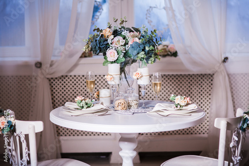 Served table for two with bouquet of spring flowers. Interior design of white room with beautiful decor. Big windows at the background. Frontal.