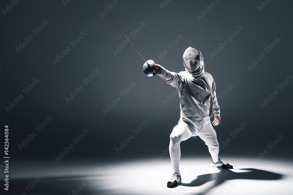 Professional fencer in fencing mask with rapier standing in position on grey Professional fencer in fencing mask with rapier standing in position on grey