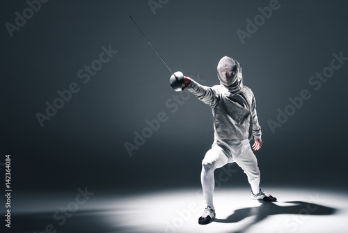Professional fencer in fencing mask with rapier standing in position on grey