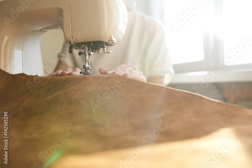 Female tailor stiching brown leather on sewing machine, close up photo