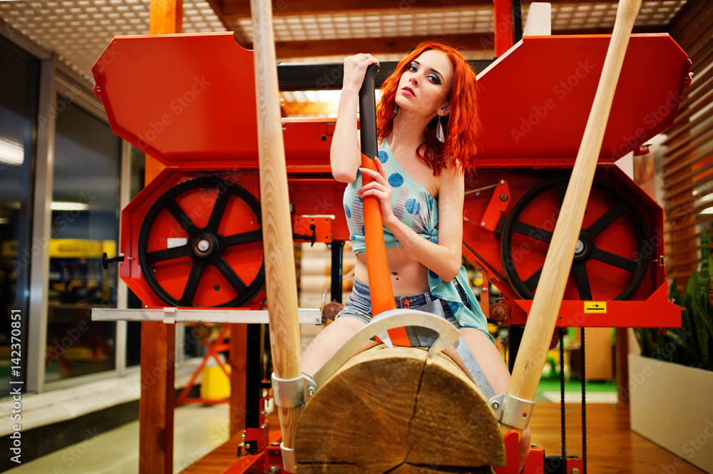 Red haired model sitting on wood sawmill with axe at store or household shop of working tools.