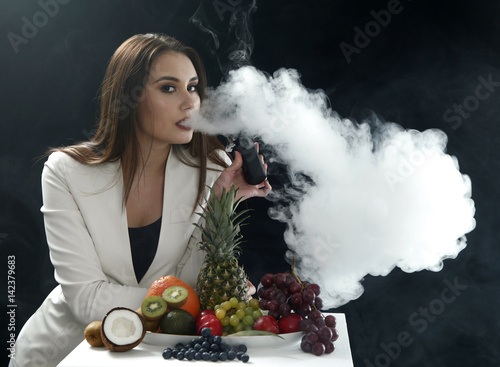 Young sexy girl smoking the vaping e cigarette close to assorti of differen fruits on black background
