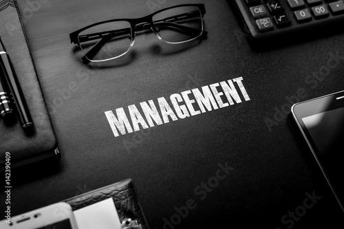 Black chalkboard with business accessories (notepad, magnifying glass, fountain pen, tablet, phone, glasses and calculator) and text MANAGEMENT. Top view.