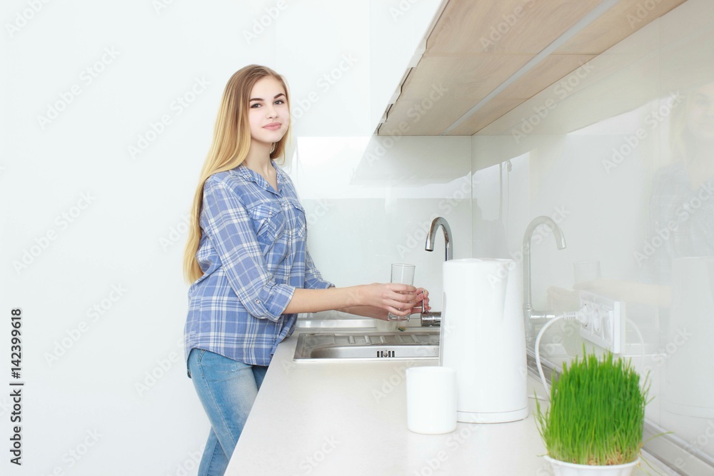 Young beautiful girl in a blue checkered shirt and jeans in the kitchen pouring the glass of water. Morning. home interior. Young beautiful girl in a blue checkered shirt and jeans in the kitchen pouring the glass of water. Morning. home interior.