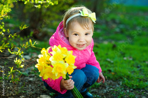 Girl With a bow on his hair sitting on the grass and holding a bouquet of yellow daffodils. A child walks outside in warm weather.