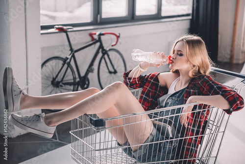 Fototapeta Young woman sitting in shopping cart and drinking water