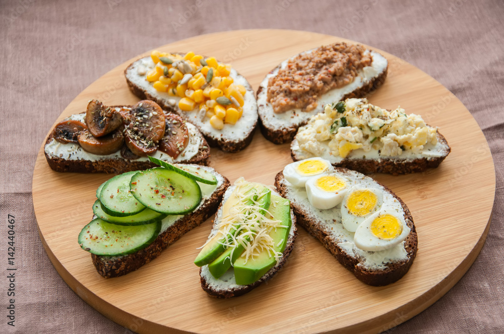 Mix of rye bread bruschettas with various filling. Sweet corn Avocado Mushrooms Quail Eggs Cucumber Omlette Tuna Seeds Cheese sandwiches served on a wooden round board. Flat lay. Mix of rye bread bruschettas with various filling. Sweet corn Avocado Mushrooms Quail Eggs Cucumber Omlette Tuna Seeds Cheese sandwiches served on a wooden round board. Flat lay.