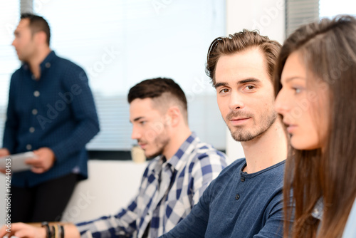 portrait of a handsome young man in high school classroom working in computer on row with teacher and classmates in background