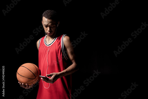 Young basketball ball in earphones holding ball and using smartphone