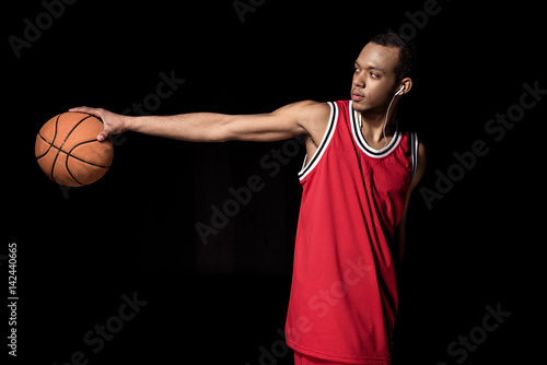Young athletic basketball player in earphones holding ball on black