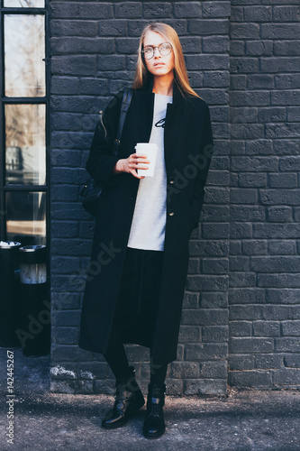 A hipster casual girl with long blond hair wearing a stylish coat and round glasses while standing on a black brick background on a street. Fashion. Style. photo