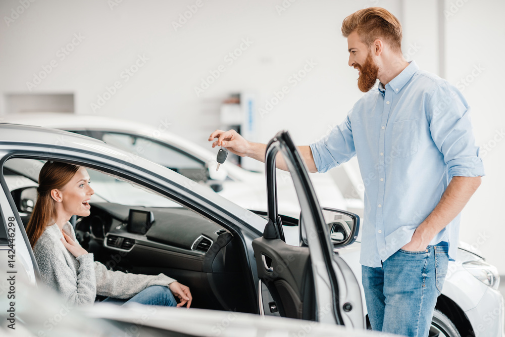 Man giving car key to woman sitting in car in dealership salon Man giving car key to woman sitting in car in dealership salon