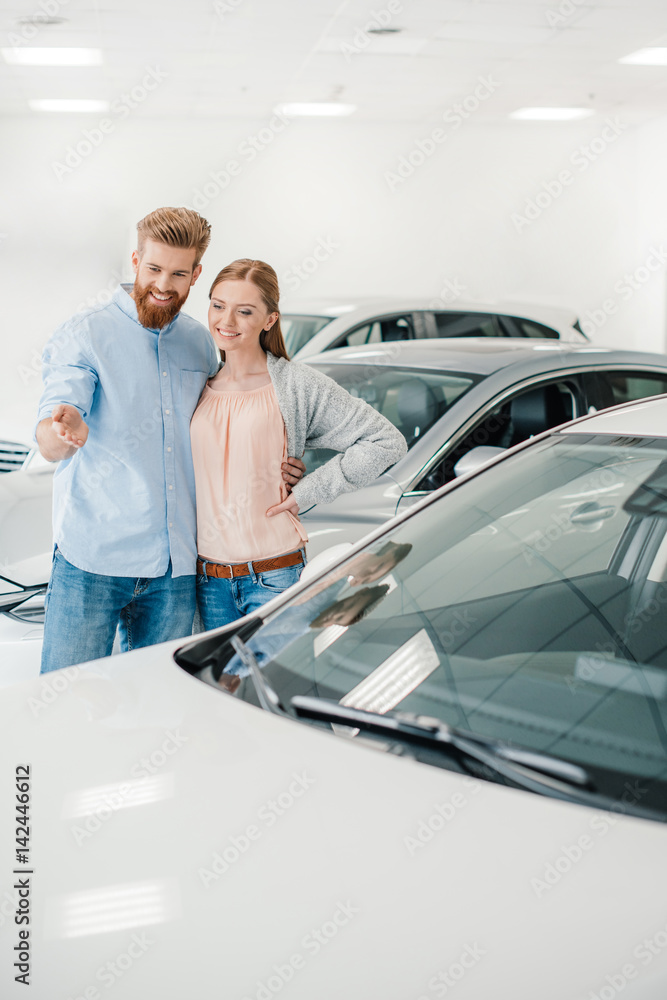 Happy couple choosing car in dealership salon, man pointing on car Happy couple choosing car in dealership salon, man pointing on car