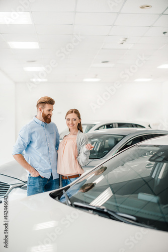 Happy couple choosing car in dealership salon, woman pointing on car