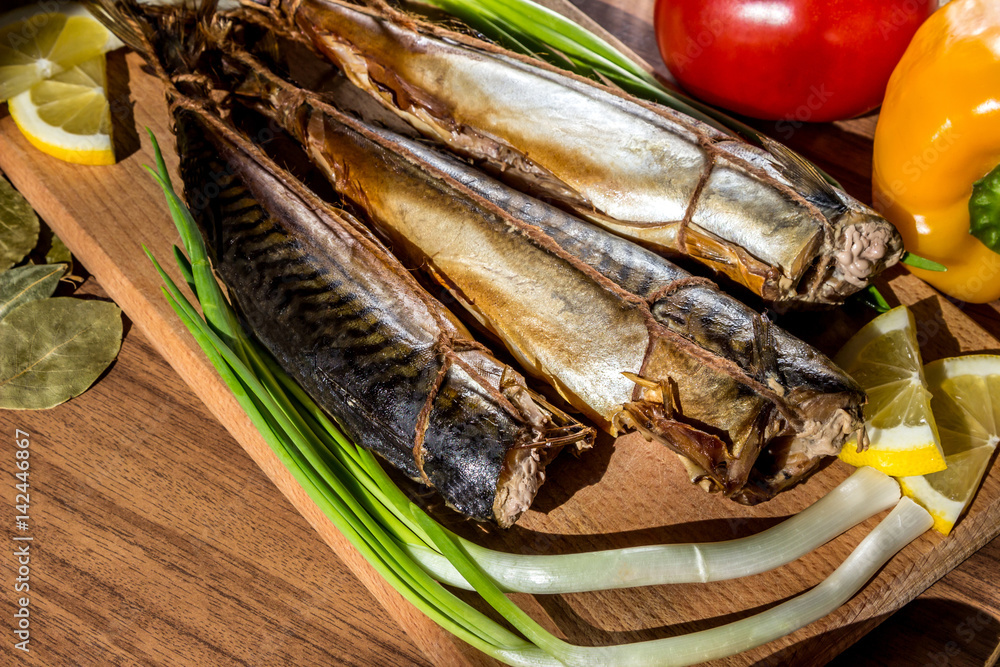 Smoked fish mackerel on wooden cutting board on background of vegetables (pepper, tomato, green onions) Smoked fish mackerel on wooden cutting board on background of vegetables (pepper, tomato, green onions)