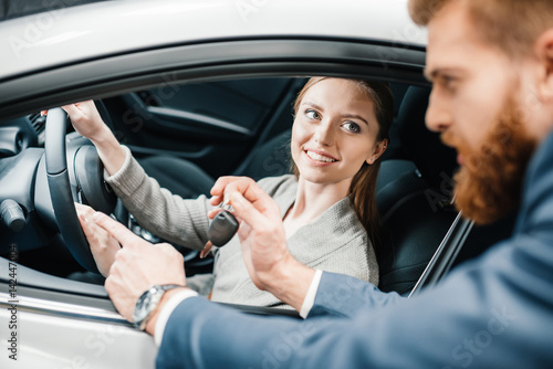 Bearded salesman giving car key to smiling young woman sitting in new car