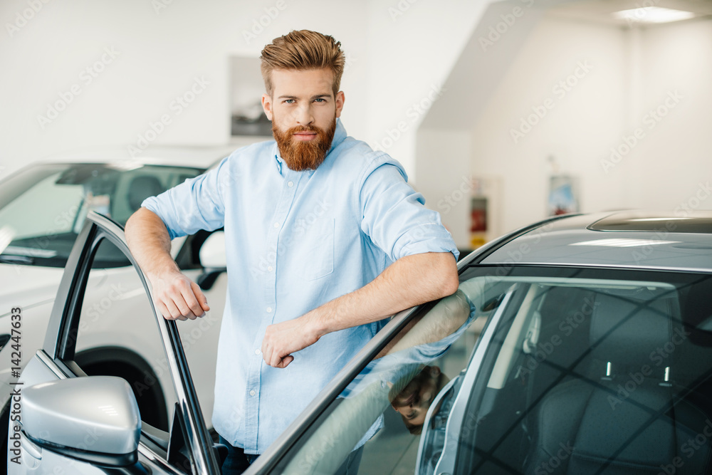 Handsome bearded young man standing with new car in auto salon Handsome bearded young man standing with new car in auto salon