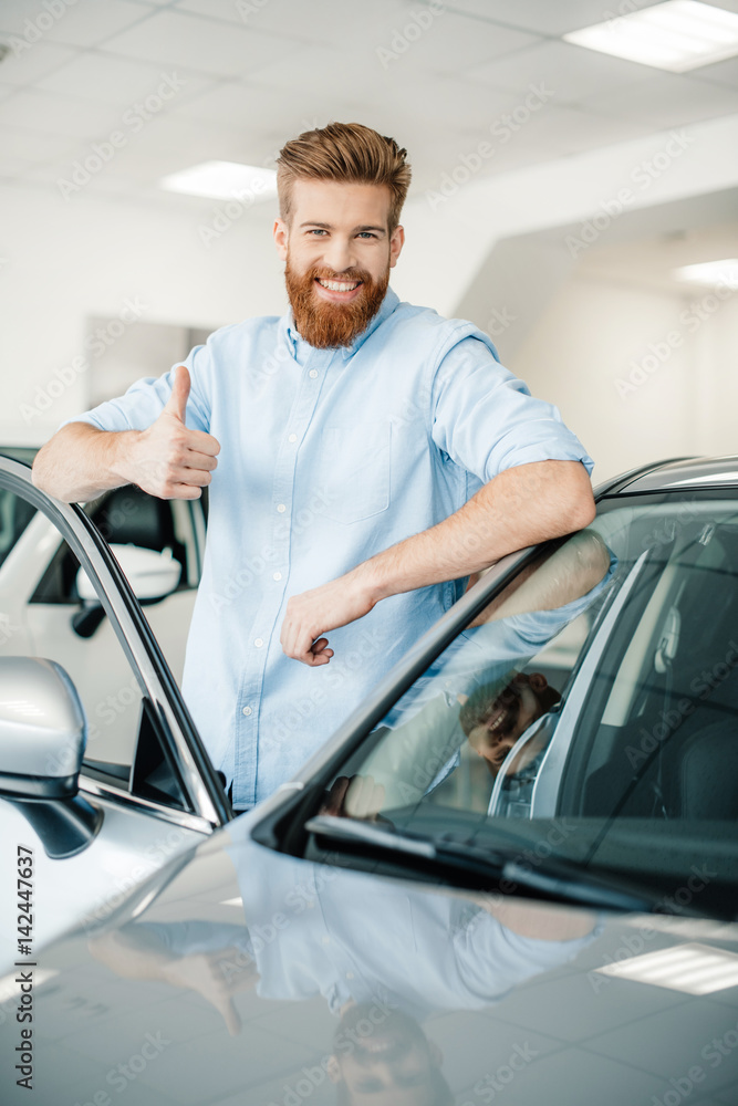 Happy young man leaning at new car and showing thumb up
