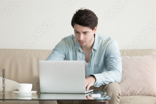 Young handsome man working online at home office using laptop computer, chatting, typing, browsing internet, checking e-mail. Distance work, relationship, self-employed, freelancer lifestyle concept
