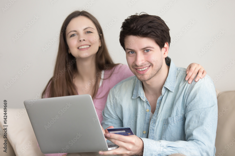 Portrait of young happy couple looking at camera, shopping online via laptop, using e-commerce, internet financial services. Man holding credit card, paying and buying, girlfriend choosing, ordering Portrait of young happy couple looking at camera, shopping online via laptop, using e-commerce, internet financial services. Man holding credit card, paying and buying, girlfriend choosing, ordering