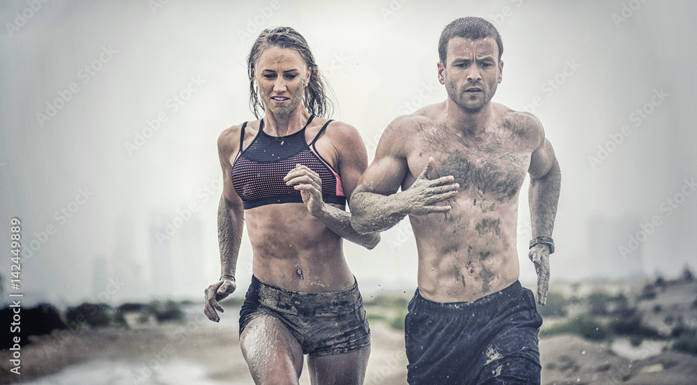 Muscular male and female athlete covered in mud running down a rough terrain with a desert background in an extreme sport race  - obrazy, fototapety, plakaty 