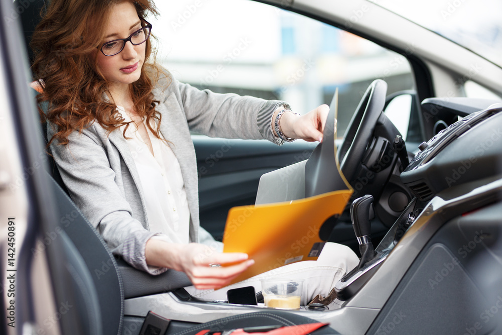 Beautiful female sales manager using laptop and phone in the car.Preparing and looking into contract papers.