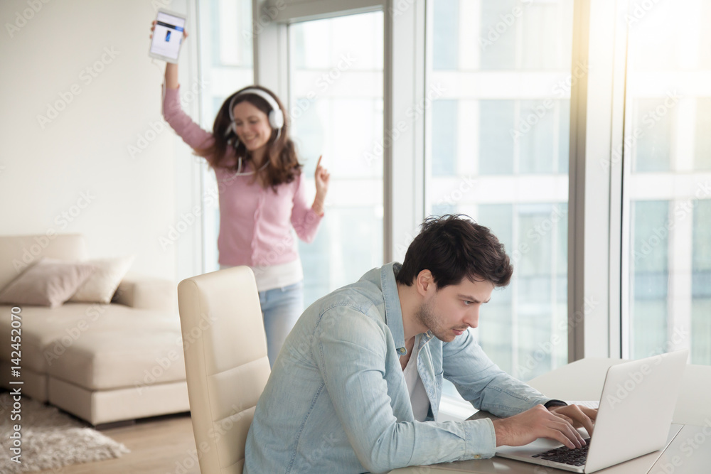 Serious man working using laptop computer, sitting at desk at home, happy relaxed woman dancing, listening to music on tablet, couple losing communication, disparate interests, ignoring each other Serious man working using laptop computer, sitting at desk at home, happy relaxed woman dancing, listening to music on tablet, couple losing communication, disparate interests, ignoring each other