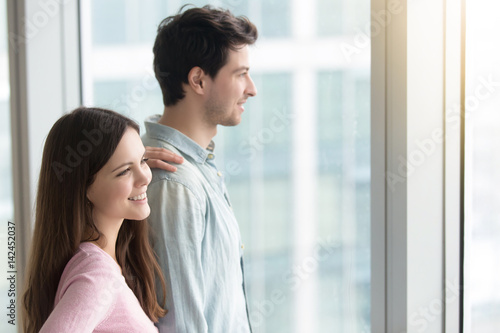 Young smiling couple looking through window at big city scenery at apartment or office, pensive man and beautiful woman dreaming and planning future, real estate purchasing, renting flat