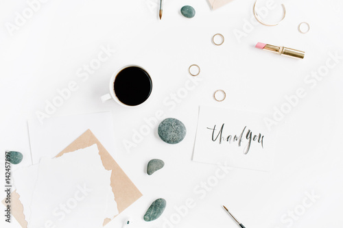Top view office table desk. "Thank you" written with calligraphic style. Workspace with coffee, craft envelope, stones, female accessories and paper blank on white background. Flat lay.