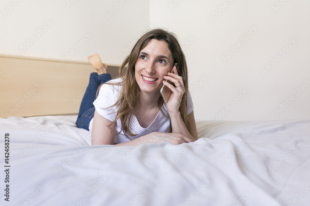 happy beautiful young woman lying on bed, talking on mobile phone and smiling. Daytime happy beautiful young woman lying on bed, talking on mobile phone and smiling. Daytime