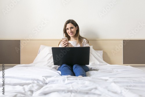 happy beautiful young woman sitting on bed using laptop at home. Daytime