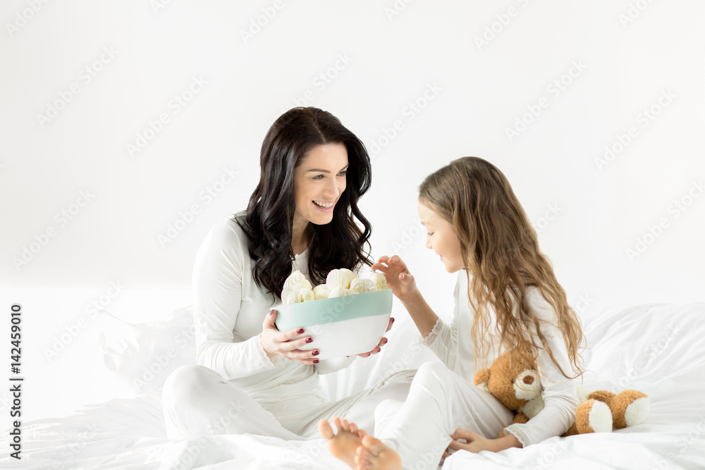 Adorable daughter and mother sitting on bed with marshmallows at home Adorable daughter and mother sitting on bed with marshmallows at home