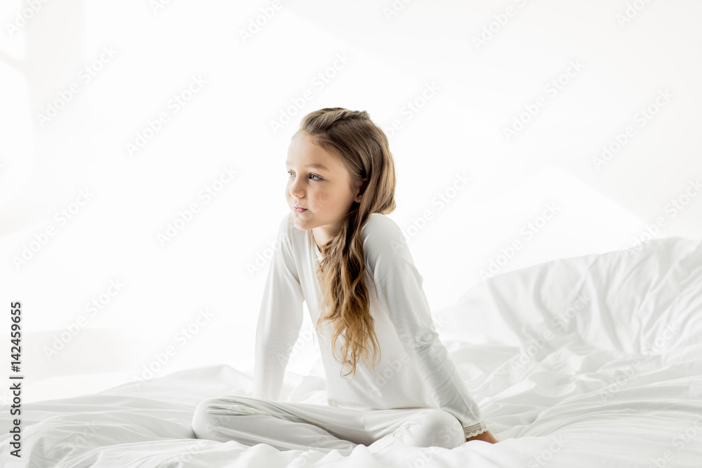 Adorable little girl in white sleepwear sitting on bed at home Stock