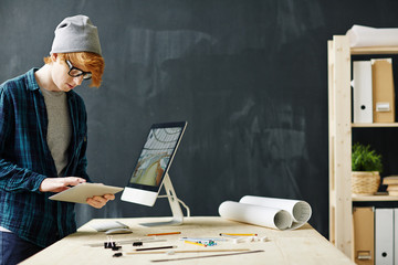 Side view portrait of young red haired architect using digital tablet standing at workplace with computer, blueprints and assorted supplies against blackboard background in office