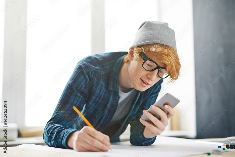 Fototapeta premium Portrait of young creative red haired man leaning on desk and using smartphone, browsing internet, while drawing with pencil on paper
