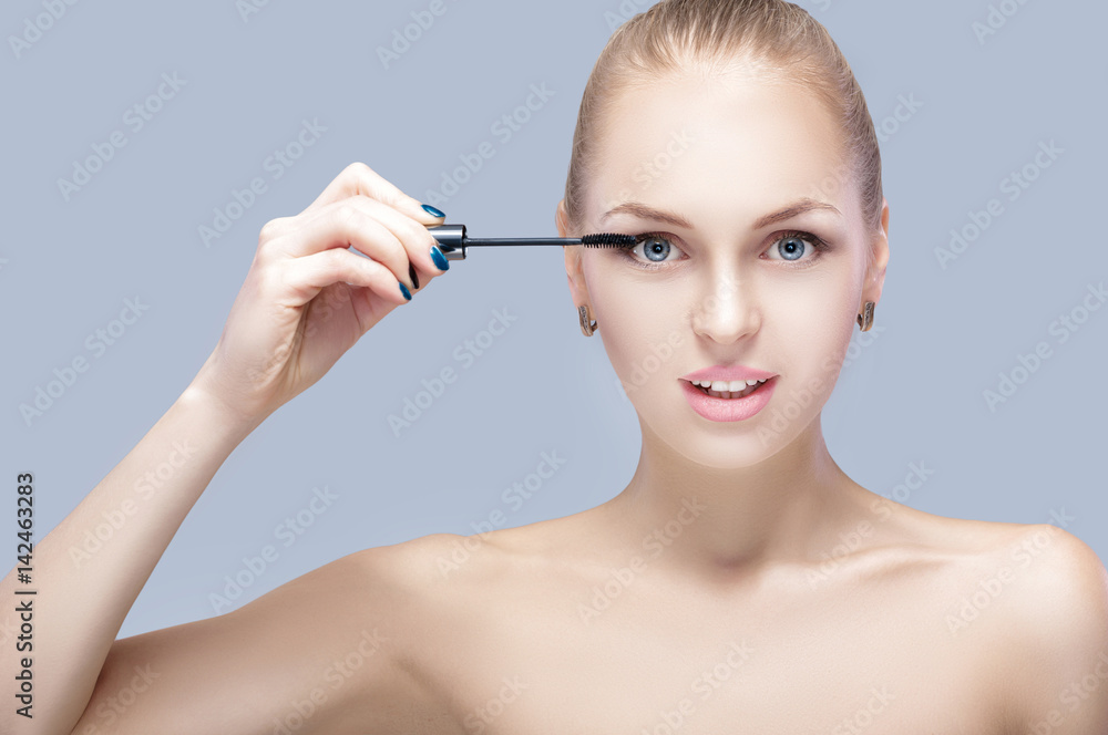 beautiful blond woman applying makeup on face on gray background. perfect makeup.  brush of mascara 