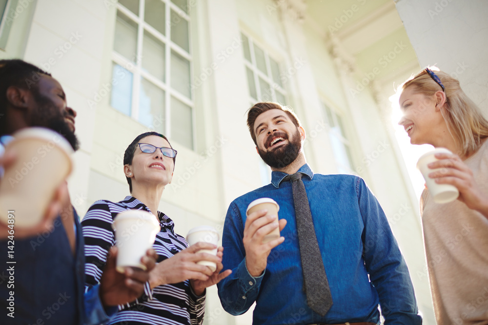 Waist-up portrait of cheerful young businessman standing outdoors with paper cup of coffee and entertaining colleagues with small talk, low angle view Waist-up portrait of cheerful young businessman standing outdoors with paper cup of coffee and entertaining colleagues with small talk, low angle view