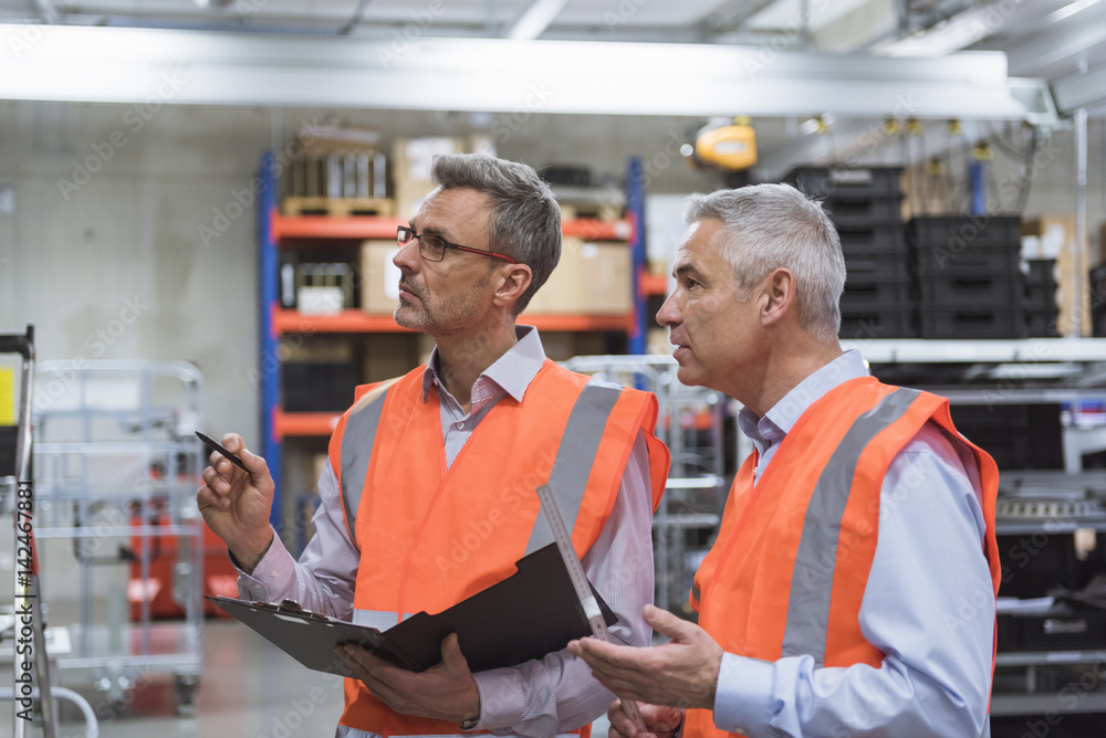 Two colleagues in factory hall wearing safety vests holding clipboard Two colleagues in factory hall wearing safety vests holding clipboard