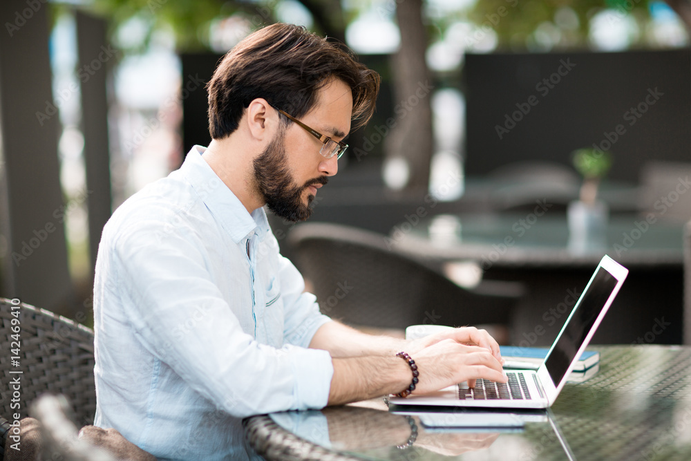 Profile view of bearded Asian employee with rolled up shirt sleeves sitting in cozy outdoor cafe and surfing Internet in order to find out necessary information, waist-up portrait Profile view of bearded Asian employee with rolled up shirt sleeves sitting in cozy outdoor cafe and surfing Internet in order to find out necessary information, waist-up portrait