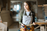 Portrait of thoughtful pretty carpenter with tool belt holding tape measure in hands and looking away in spacious workshop