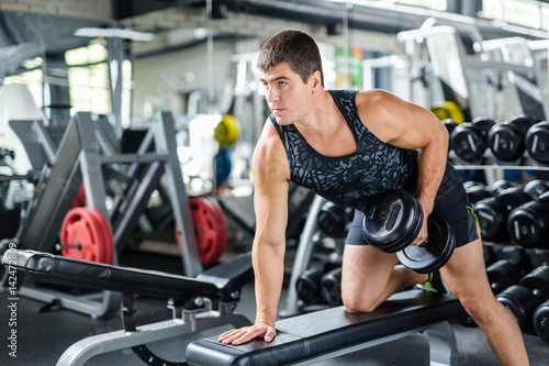 Portrait of muscular man working out with dumbbells on bench in modern gym, flexing and pumping arm muscles