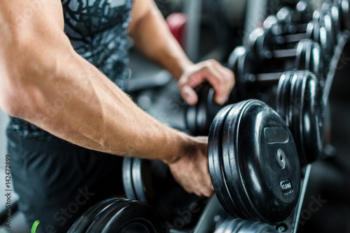 Closeup shot of muscular male arm picking up heavy dumbbell weights from equipment rack in modern gym
