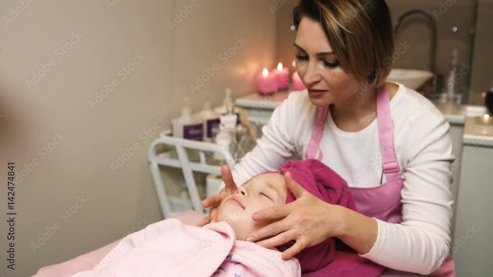 Professional beautician applying mask and doing massage on beautiful little girls face at beauty salon. Spa therapy. Close up view Professional beautician applying mask and doing massage on beautiful little girls face at beauty salon. Spa therapy. Close up view