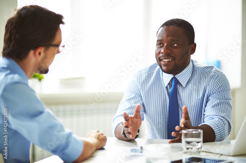 Afro-American bearded HR manager sitting at office desk while conducting interview with male applicant for position photo