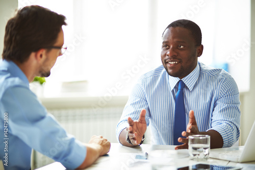 Afro-American bearded HR manager sitting at office desk while conducting interview with male applicant for position